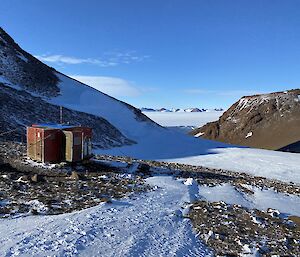 Small field hut sits in left mid picture in valley between two rocky peaks. In the distance in another rocky mountain range.