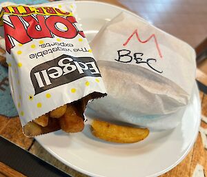 Plate sitting on wooden bench holding a burger wrapped in paper and a bay of potato wedges