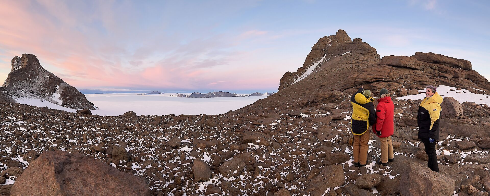 In the foreground three people in down coats standing on rough rocky terrain. Rock mountain peaks on both sides of photo. In the distance the ice plateau highlighted by sunset.
