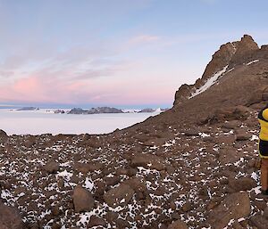 In the foreground three people in down coats standing on rough rocky terrain. Rock mountain peaks on both sides of photo. In the distance the ice plateau highlighted by sunset.