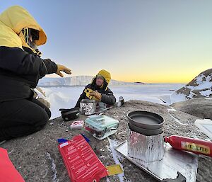 Camp stoves set up on large rock in foreground of picture. Two expeditioners sit to side of rock wearing large down jackets with hoods raised. In the background ice covered bay with ice cliffs at sunset.