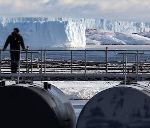 In foreground man standing on walkway on top of two large fuel tanks, in background two large tabular icebergs