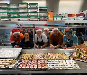 A group of people in Santa hats with trays of Christmas cookies in a large kitchen
