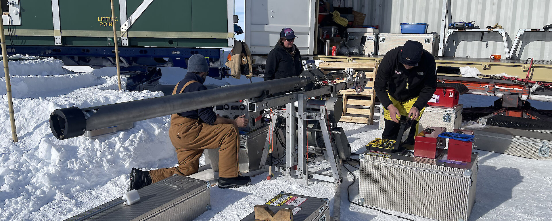 Three people out on the snow in front of a large, open transport container containing assorted trunks, toolboxes and other gear. They are examining some drilling apparatus, a slim cylindrical device a few metres long, mounted on a metal stand in a horizontal position