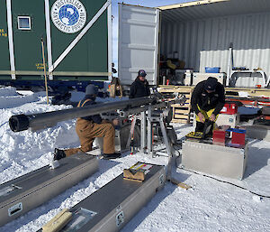 Three people out on the snow in front of a large, open transport container containing assorted trunks, toolboxes and other gear. They are examining some drilling apparatus, a slim cylindrical device a few metres long, mounted on a metal stand in a horizontal position