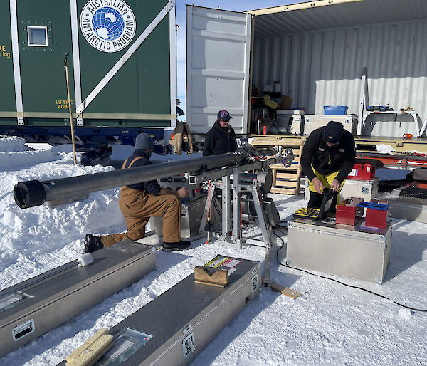 Three people out on the snow in front of a large, open transport container containing assorted trunks, toolboxes and other gear. They are examining some drilling apparatus, a slim cylindrical device a few metres long, mounted on a metal stand in a horizontal position