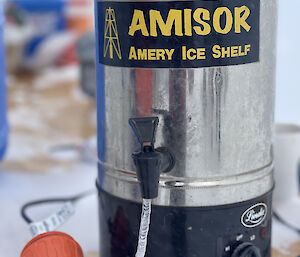 A close-up photo of a tea-making urn on an outdoor table - evidently a very cold location, as there are patches of snow on the table, and a small icicle coming out of the urn's tap