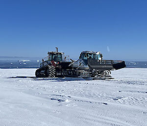 A tractor is towing another similar tracked vehicle, which has been mounted on large skis for easy transport across the snow-covered ground