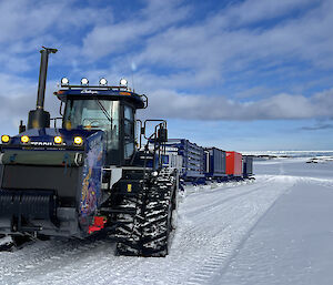 A front view of a tractor which is towing a train of 4 sled-mounted transport containers, like a caravan, on a groomed track across a snowy plain