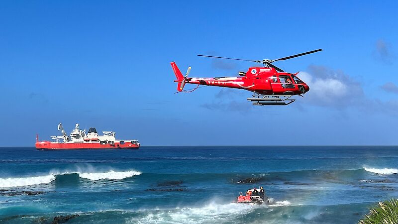 A helicopter hovers over the water with a red ship in the background