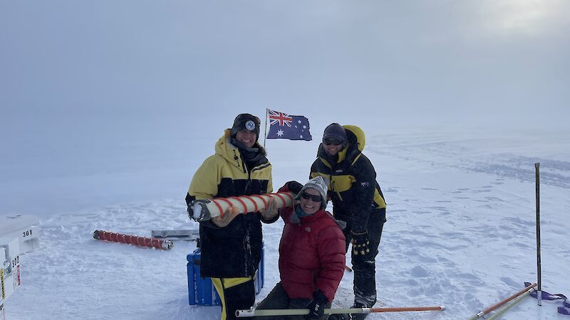 Three people hold an ice core surrounding by ice with an Australian flag behind them