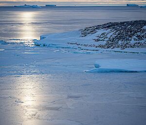 An ocean scene showing a large swathe of new, thin sea ice, formed in a patchwork manner. There are large icebergs on the horizon. A setting sun shining through clouds casts a dull silvery sheen on ice and water