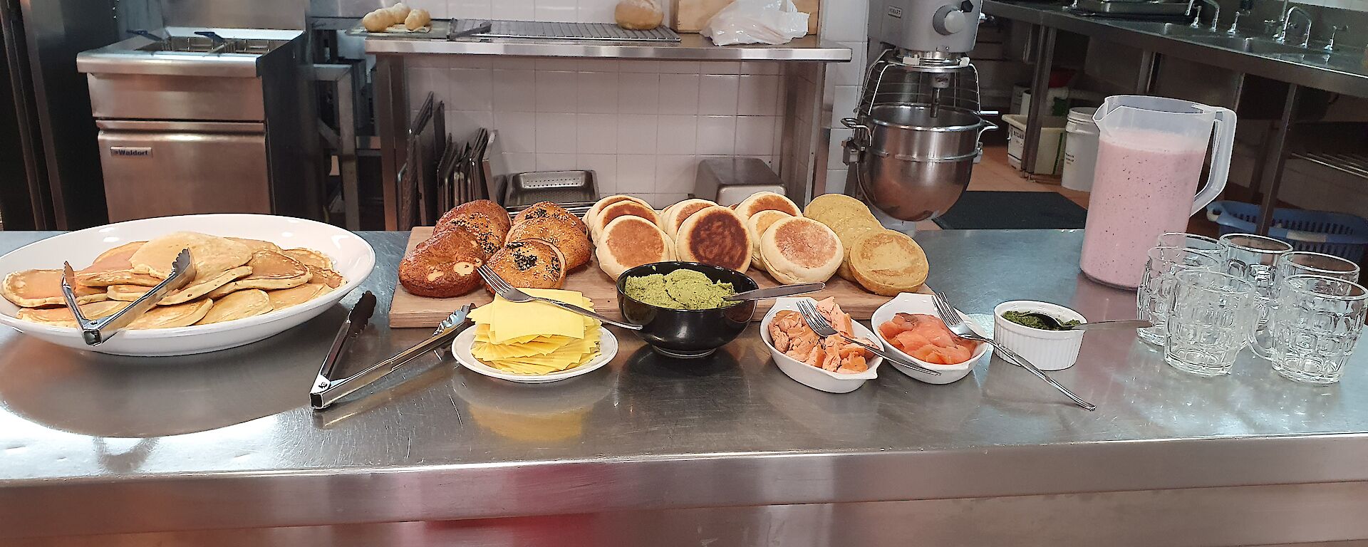 Food laid out on kitchen bench, from left to right, platter of pancakes, wooden chopping board with bagels, english muffins and crumpets with accompaniments in front (cheese, avocado, smoked salmon, herbs), and jug of berry smoothie with glasses