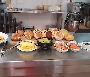 Food laid out on kitchen bench, from left to right, platter of pancakes, wooden chopping board with bagels, english muffins and crumpets with accompaniments in front (cheese, avocado, smoked salmon, herbs), and jug of berry smoothie with glasses