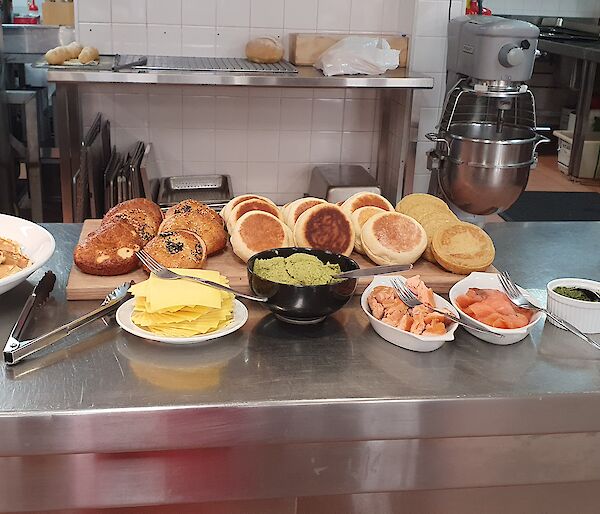 Food laid out on kitchen bench, from left to right, platter of pancakes, wooden chopping board with bagels, english muffins and crumpets with accompaniments in front (cheese, avocado, smoked salmon, herbs), and jug of berry smoothie with glasses