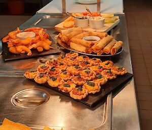 Five trays of food set out on kitchen bench, closest to furthest away, corn chips and dips, mini quiches, spring rolls, crumbed prawns, and dips and biscuits