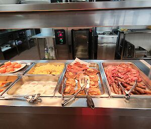 Buffet of brunch items in a bein-marie - bacon, sausages, corn fritters, tomatoes, egg scrambled and poached, backed bean and spaghetti