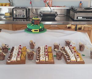 Two teired table covered with white tablecloth, on bottom tier four trays with rows of four different desserts and on the top teir a cheese board on left and in centre a two teir cake covered in green icing with irish decorations