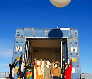 Met balloon is launched and rising into the air, below are a group of expeditioners in a variety of onesies looking up at the balloon and standing in front of the station balloon building which is square, blue and has large doors which open to allow the release of the balloon.