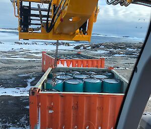 Open topped orange 1/4 height shipping container filled with blue fuel drums, JCB telehandler arm is extended over the fuel drums and attached via a drum lifter to one drum