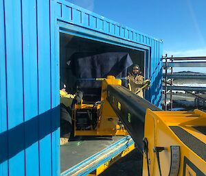 Blue shipping container with hole in side wall, two men stand inside, camera view is down the length of the telehandler's arm into the building to the freezer at the end.