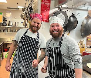 Two men wearing stripped aprons and bandanas smile at camera and each hold a knife in their hands