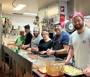 Six expeditioners in aprons and bandanas stand along a kitchen bench which is laden with pizza trays and containers of pizza toppings.