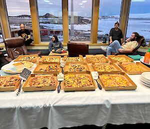 In the foreground a large table which is covered in pizzas in open pizza boxes, in background people lounging in arm chairs in front of windows with view of ice covered bay outside