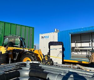 Telehandler lift a large 2 m high freezer on its forks, into a blue shipping container that has a large hole in its side.  Two men stand holding radios, directing the movement of the JCB.