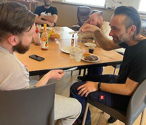 4 men sitting around a table, 2 of them with butter knives pointing at each other in jest