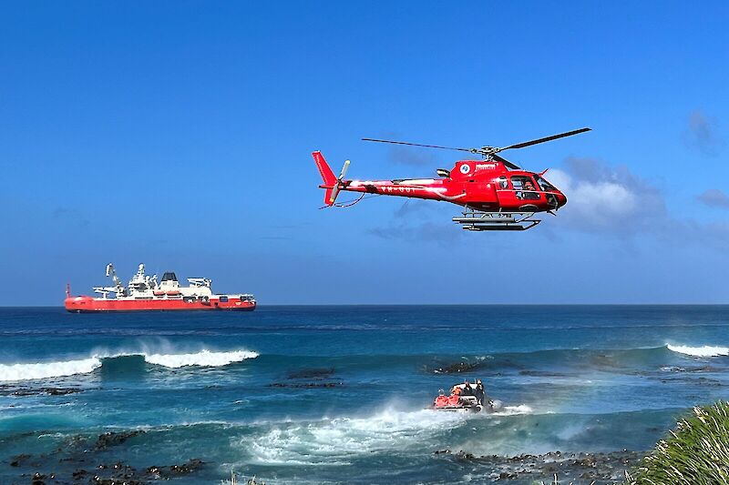 Red helicopter hovering near the shore with a LARC and the Nuyina in the background