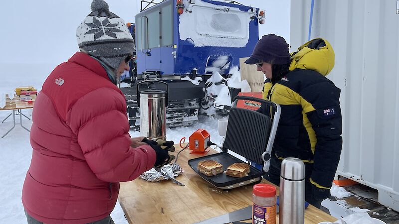 Two people in warm clothing cook toasted sandwiches surrounded by ice