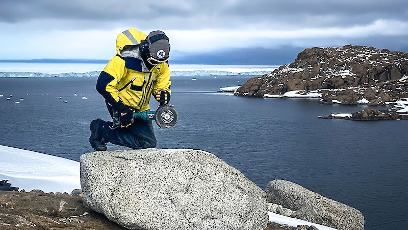 A woman leans against a rock with an angle grinder with the ocean and ice behind her