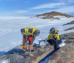 A man drills into a rock while a woman watches on. Both are on the side of a rocky hill with snow and ice behind them