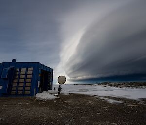 A person stands just outside the doorway of a large shed carrying a weather balloon in one hand. An enormous, layered cloud formation takes up half the sky in the background