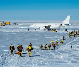 An A319 passenger plane is parked on a snowy plain. A long queue of people wearing yellow and black weatherproof clothing and carrying hand luggage is walking towards the stairs leading up to the plane's rear door