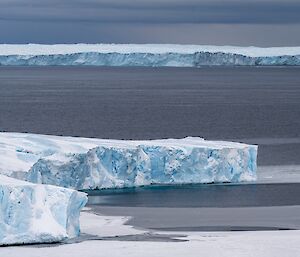 The edges of a glacier, appearing as craggy shelves of ice marbled blue and white, abutting a steel-grey ocean