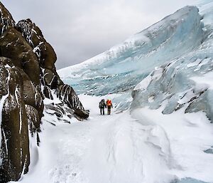 Two people walking through a wind scour - a channel carved by wind over the ages, like a snow-lined gorge with a rock wall on one side and an ice wall on the other