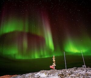 The Aurora Australis in the sky above a signpost and flagpoles. It appears as ribbons of green light, with maroon upper edges fading flame-like into the night