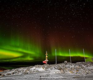 The Aurora Australis in the sky above a signpost, flagpoles and a shed. It appears like lakes and bands of green light, fading through dull red on their upper edges into a starry night sky
