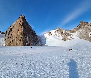 Expeditioners in the distance walking across blue ice which large fang shaped rocky mountain looms above