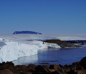 Ice covered bay with ice cliffs rising above and then leading to the ice plateau, blue skies above and in the distance a rocky  mountain range