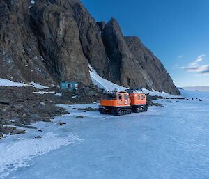 Ice plateau abuts rocky mountain range, where they meet is a small blue field hut and an orange hagglunds parked in front