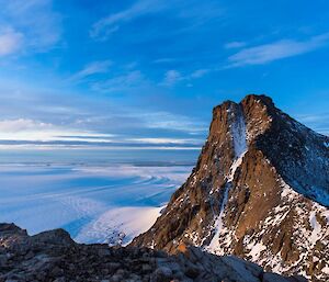 In foreground rocky mountainous peak then looking across ice plateau to the ocean, blue skies with whispy clouds above