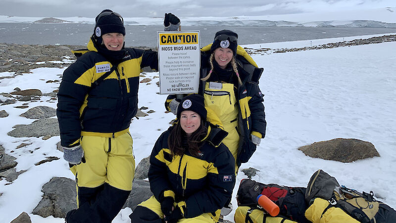Three scientists in yellow survival clothing stand next to a sign that cautions people about fragile moss beds