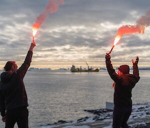 Two people holding flares with a ship departing in the background