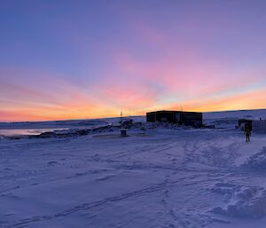 A large shed seen across a wide, snowy road crossed with many tracks and footprints. In the dawn shadow the snow appears dim violet-blue. Thin streaks of cloud reach across the sky, lit up in pink and fiery gold by the sun from below the horizon