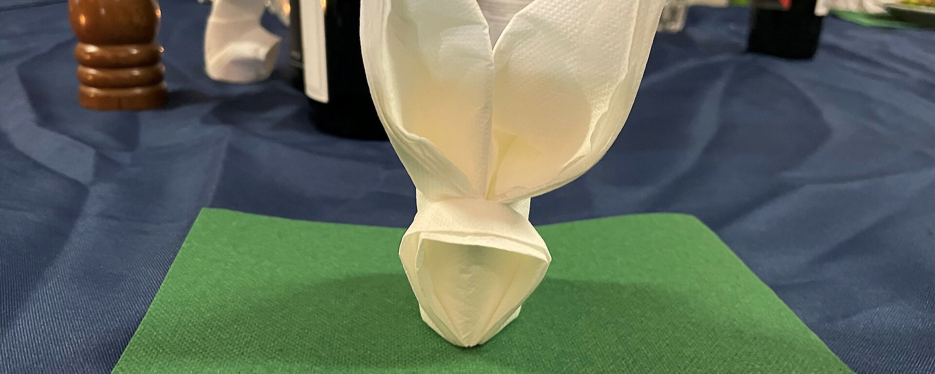 A white paper napkin folded into the shape of a rabbit's head, placed on a dining table in front of some wine bottles