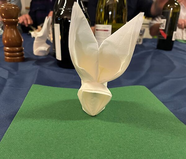 A white paper napkin folded into the shape of a rabbit's head, placed on a dining table in front of some wine bottles
