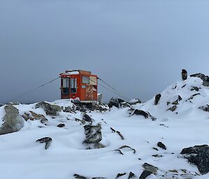 The cabin of a D4 tractor demounted from its vehicle, fitted with a wooden door and secured with guywires amidst rocks and snow